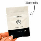 Hand holding an AEMBR Heritage laundry powder sample packet, labeled for 2 loads.