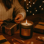 AEMBR Juletid non-toxic candle burning beside hands wrapping gifts, warm festive ambiance.