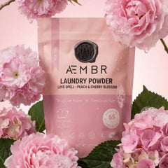 AEMBR Love Spell laundry powder in pink packaging, surrounded by cherry blossoms and green leaves.