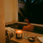 Woman enjoying a relaxing salt bath with AEMBR non-toxic candle and bath salts nearby.
